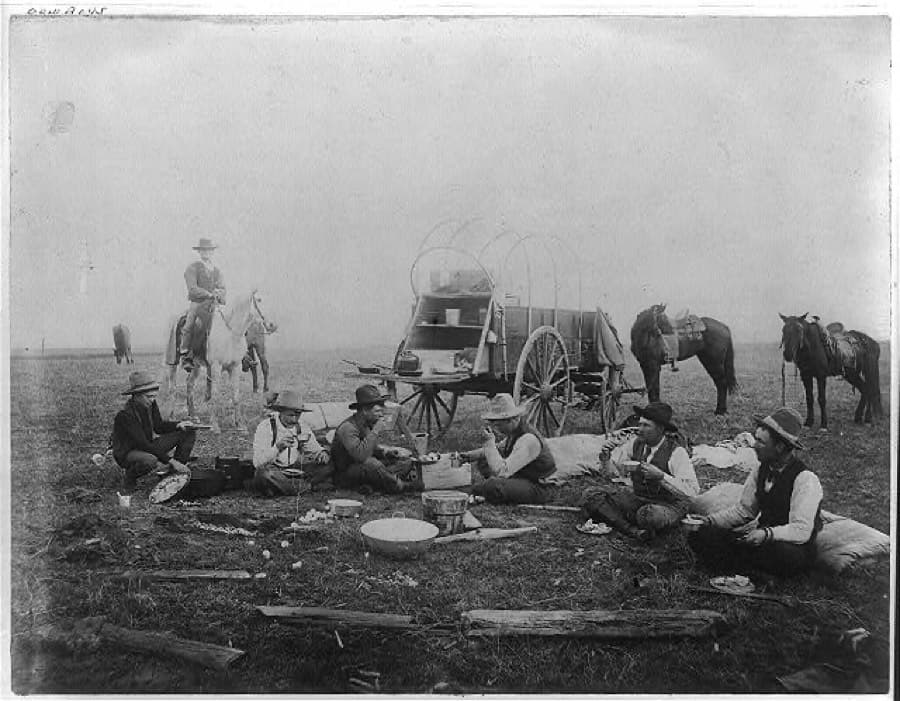 Cowboys eating on the range in 1880 with a chuck wagon behind them, its chuck box open